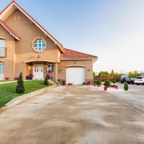 The exterior of Casa Iulia is showcased, featuring a well-maintained facade with a prominent entrance and a garage. Potted plants line the pathway, and a generous parking area is visible, accommodating multiple vehicles. The roof is adorned with red tiles under a clear blue sky.
