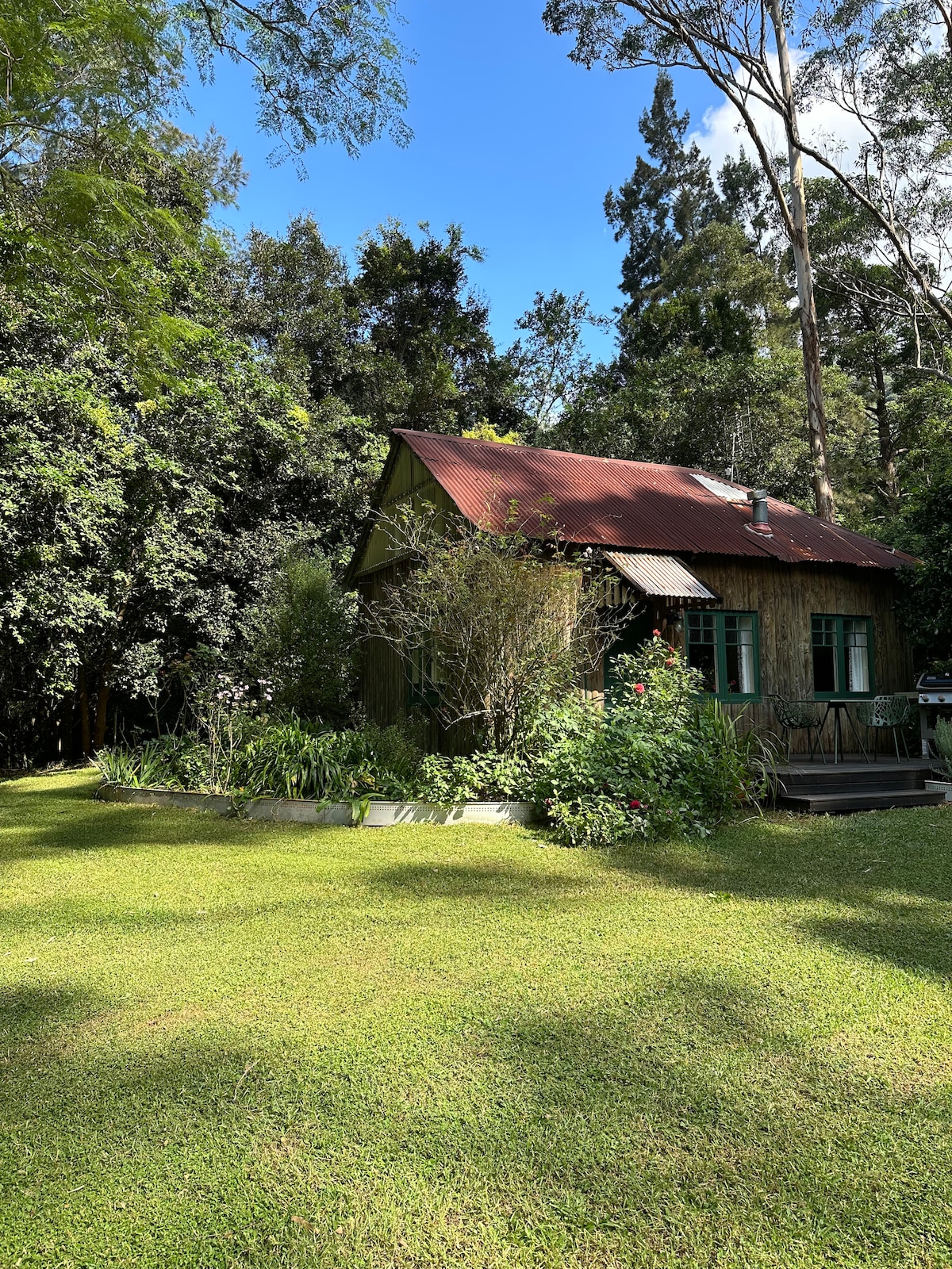 A rustic wooden cottage with a corrugated iron roof is surrounded by lush greenery. Tall trees provide shade, while the well-maintained lawn enhances the tranquil outdoor setting. Flowering plants add subtle color, creating a peaceful retreat amidst nature.