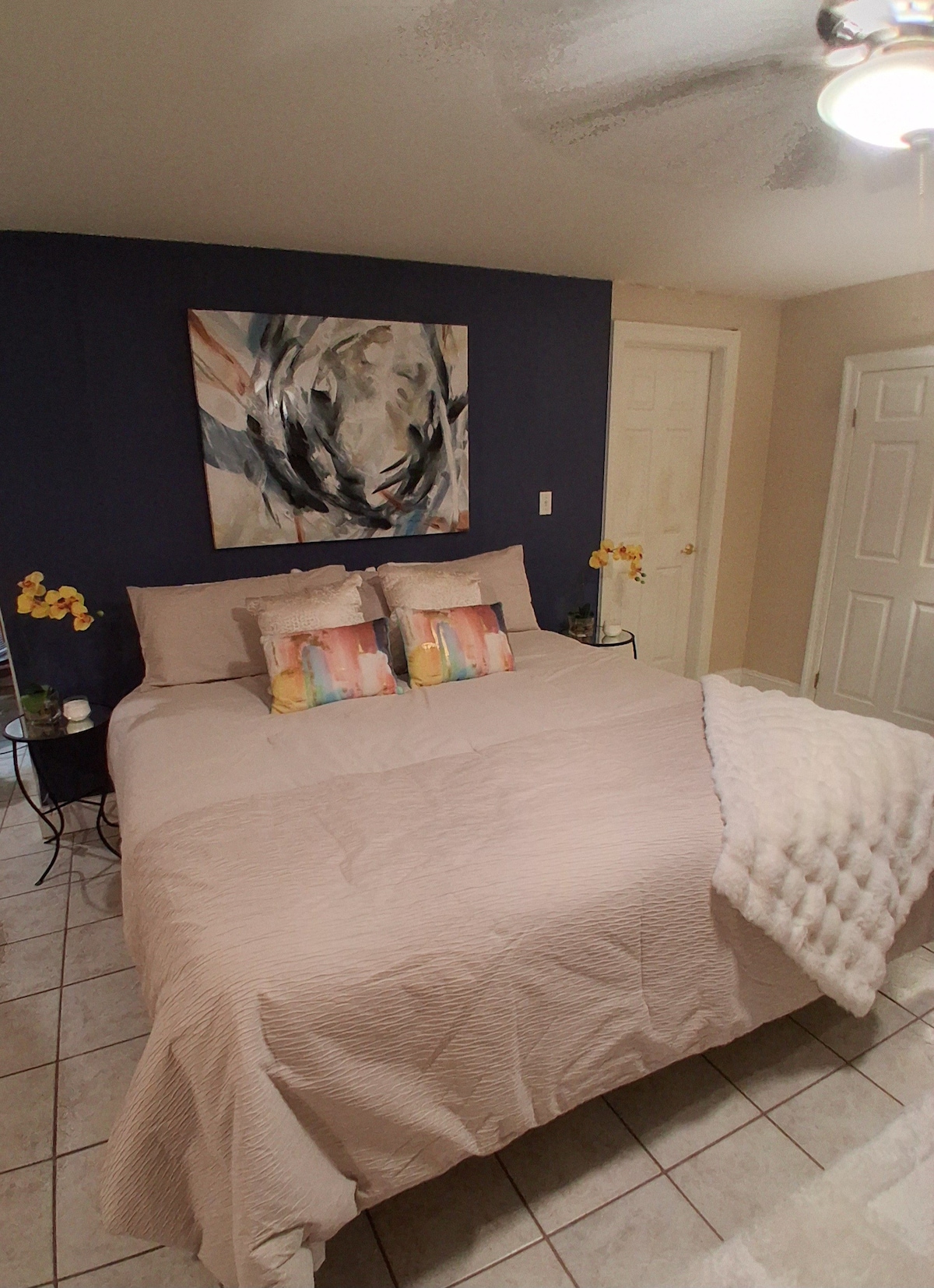 A spacious bedroom is highlighted by a plush bed adorned with decorative pillows and a soft blanket. A large piece of abstract art decorates the blue wall above, while cheerful yellow orchids add a touch of brightness to each side of the bed.
