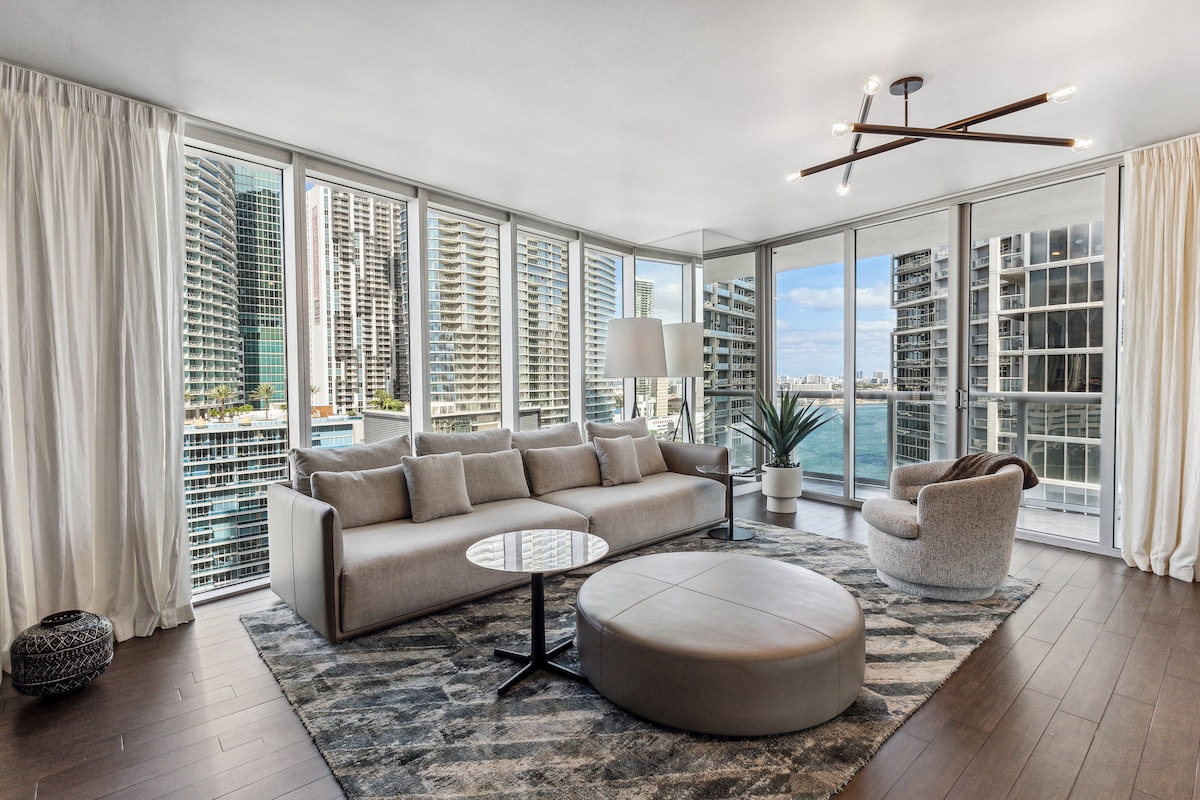 A modern living area features a spacious sofa and a circular ottoman nestled on a soft area rug. Floor-to-ceiling windows provide expansive views of the Miami River and surrounding high-rise buildings, allowing natural light to fill the room.