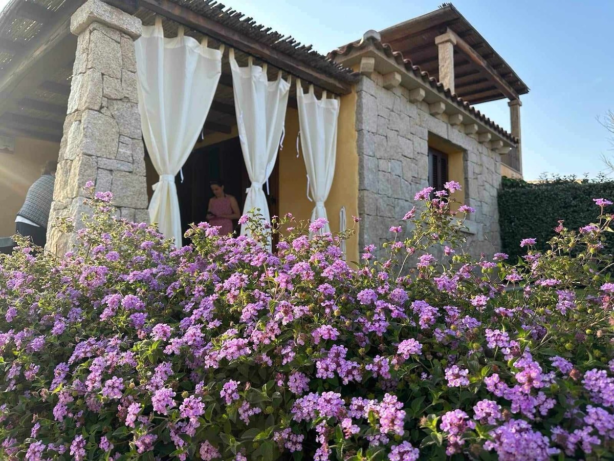 A charming stone house features soft white drapes framing the entrance. Vibrant purple flowers adorn the front garden, contributing to a serene setting. Natural wooden elements are visible on the roof, enhancing the overall rustic aesthetic of the home.