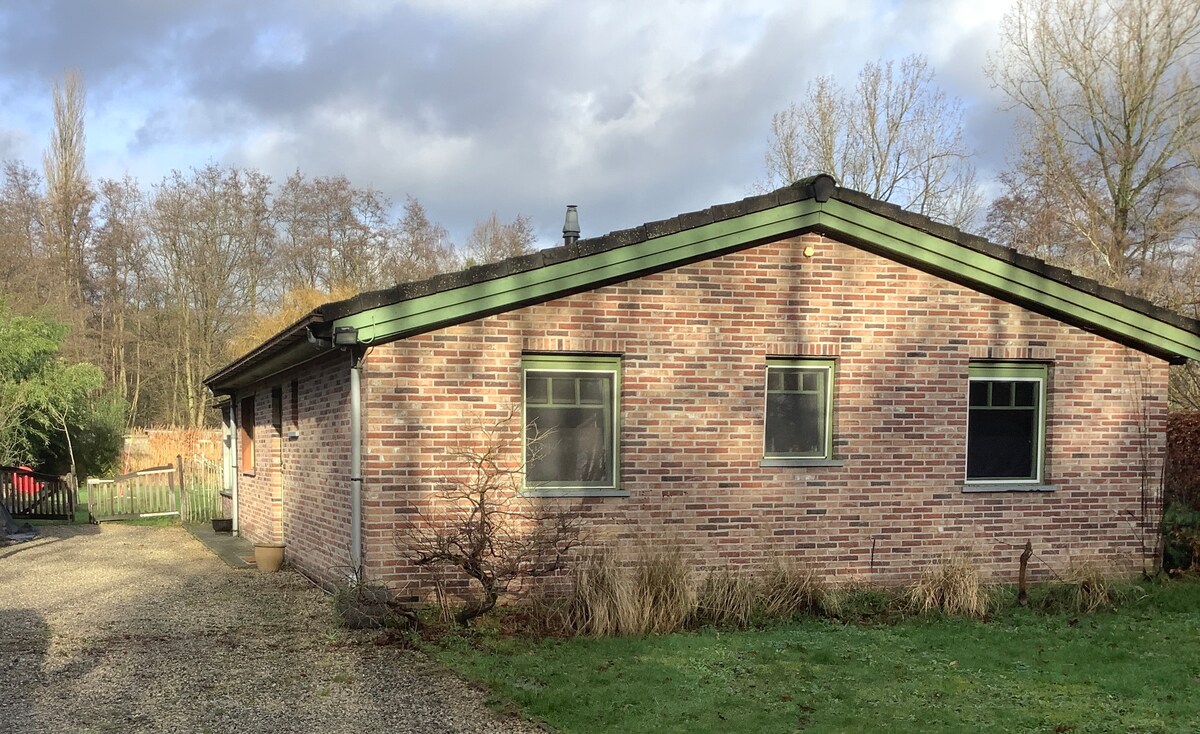 A brick bungalow is set in a natural environment, featuring large windows on the facade that allow for natural light. The property is surrounded by greenery and a gravel pathway leading to the entrance, providing a sense of tranquility.