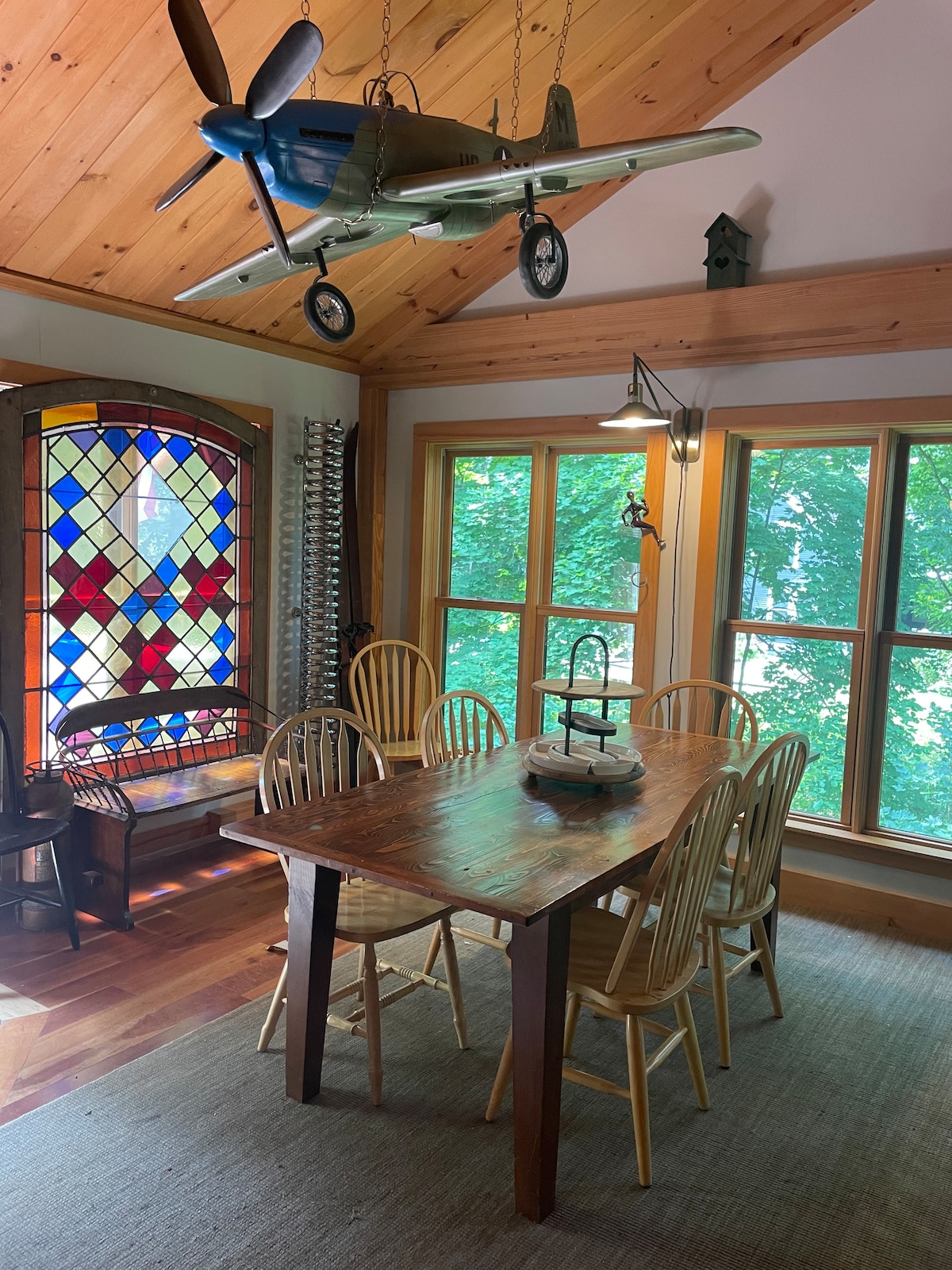 A rustic dining area features a wooden table surrounded by six chairs, under a vaulted ceiling adorned with wood beams. A stained glass window casts colorful light, and a vintage airplane model hangs above, adding a unique touch to the inviting space.