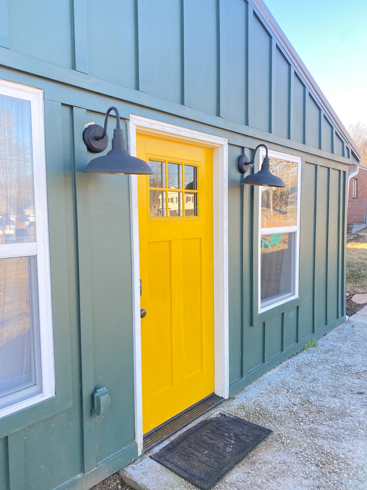 The vibrant yellow front door stands out against the dark green exterior of the building. Two black sconces flank the door, providing illumination. Nearby windows allow natural light to enhance the exterior's welcoming appearance.