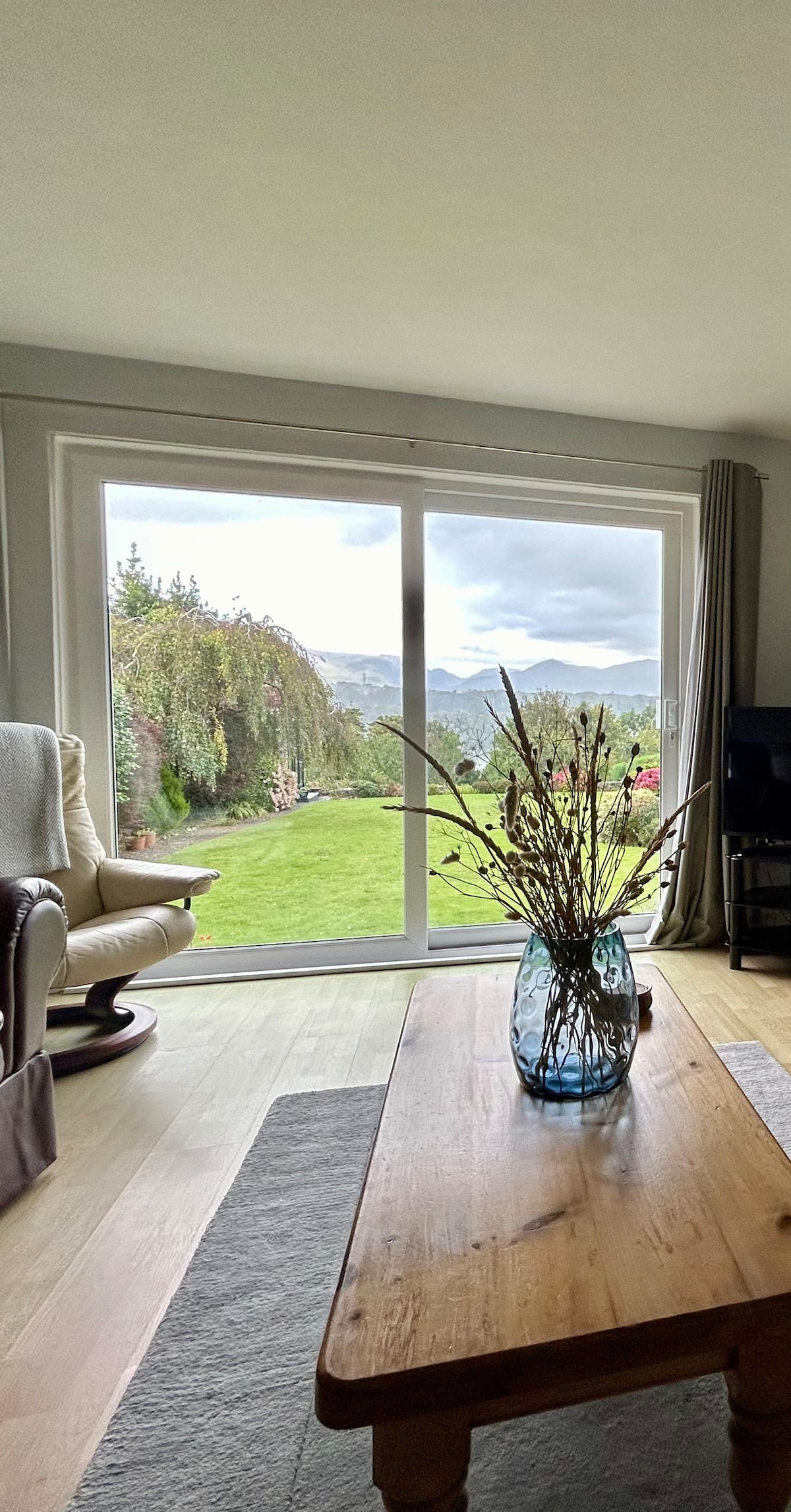 A bright living area features a large glass sliding door that opens to a view of the garden and mountains. A wood table sits in front, adorned with a blue vase holding dried flowers. A comfortable armchair is positioned nearby, creating a cozy spot to relax.