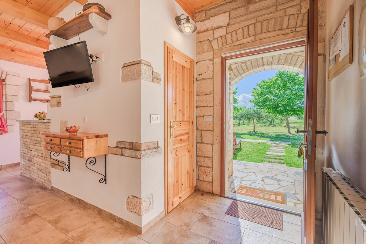 The entrance showcases a well-lit space with a wooden door leading to a stone-paved walkway. A flat-screen TV is mounted on the wall, and a wooden console with drawers provides storage. Natural light illuminates the area, creating a welcoming ambiance.