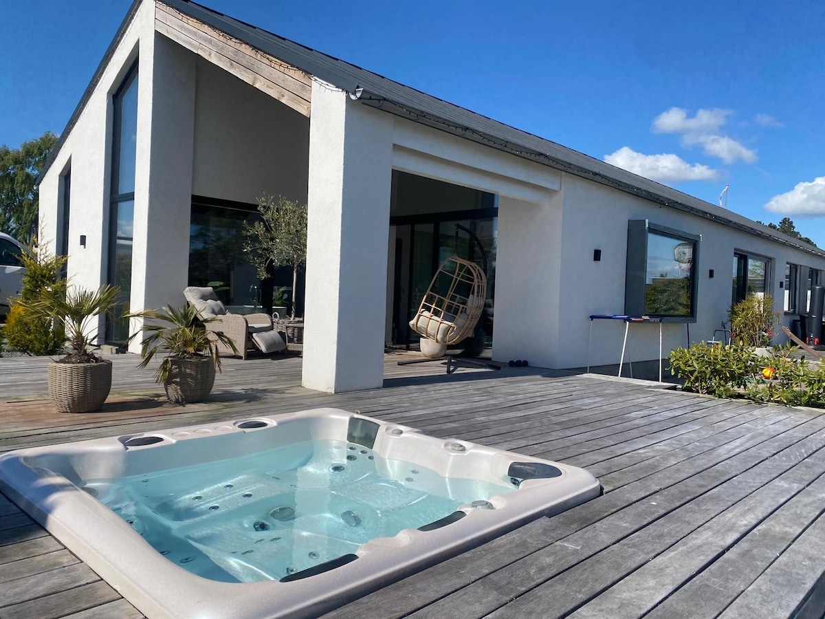 A modern house is seen from the terrace, featuring large glass windows and a stylish hanging chair. A hot tub is positioned on the wooden deck, surrounded by potted plants and garden seating, creating a relaxed outdoor atmosphere.