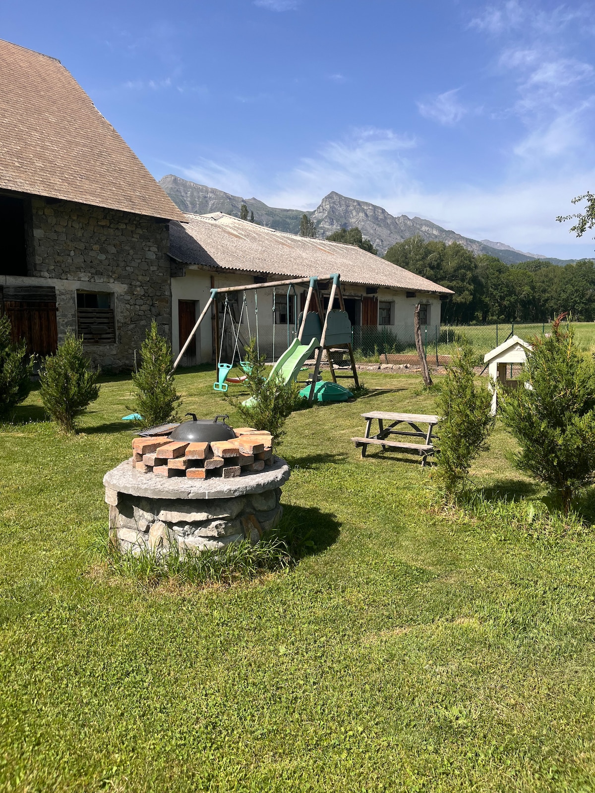 A grassy outdoor area features a stone fire pit surrounded by low bushes. A playground with swings and a slide is visible, alongside a picnic table. The background showcases a rustic building structure and mountains under a clear blue sky.