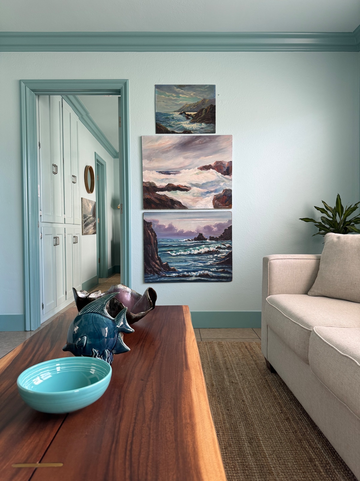 A cozy living area features soft ocean-themed artwork displayed on a light-colored wall. A wooden coffee table holds a blue bowl and sculptural decor, while a neutral-toned couch is positioned nearby. Natural light gently illuminates the space, creating a serene atmosphere.