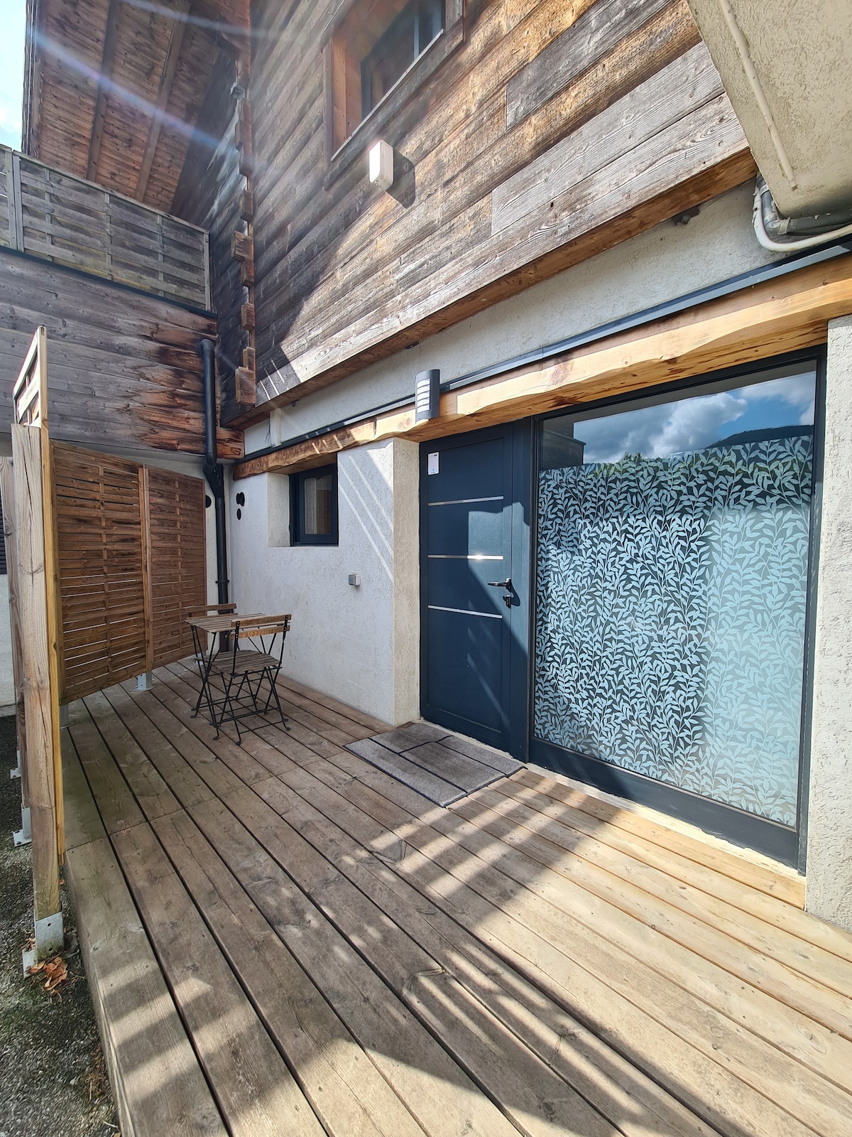 The entrance to the apartment is highlighted by a stylish door with a frosted glass panel. A small terrace area features a wooden deck and a bistro table with two chairs, surrounded by wooden fencing that offers privacy.