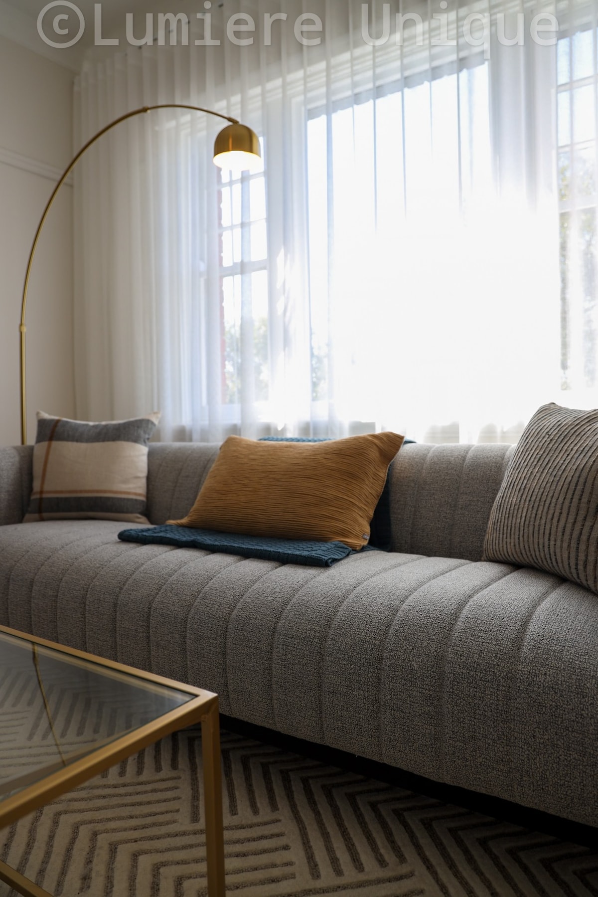 A sleek, modern sofa is positioned near large windows adorned with sheer curtains, allowing natural light to filter through. Decorative cushions in various textures enhance the sofa's appeal. A gold-framed coffee table sits in front, complemented by a patterned area rug underneath.