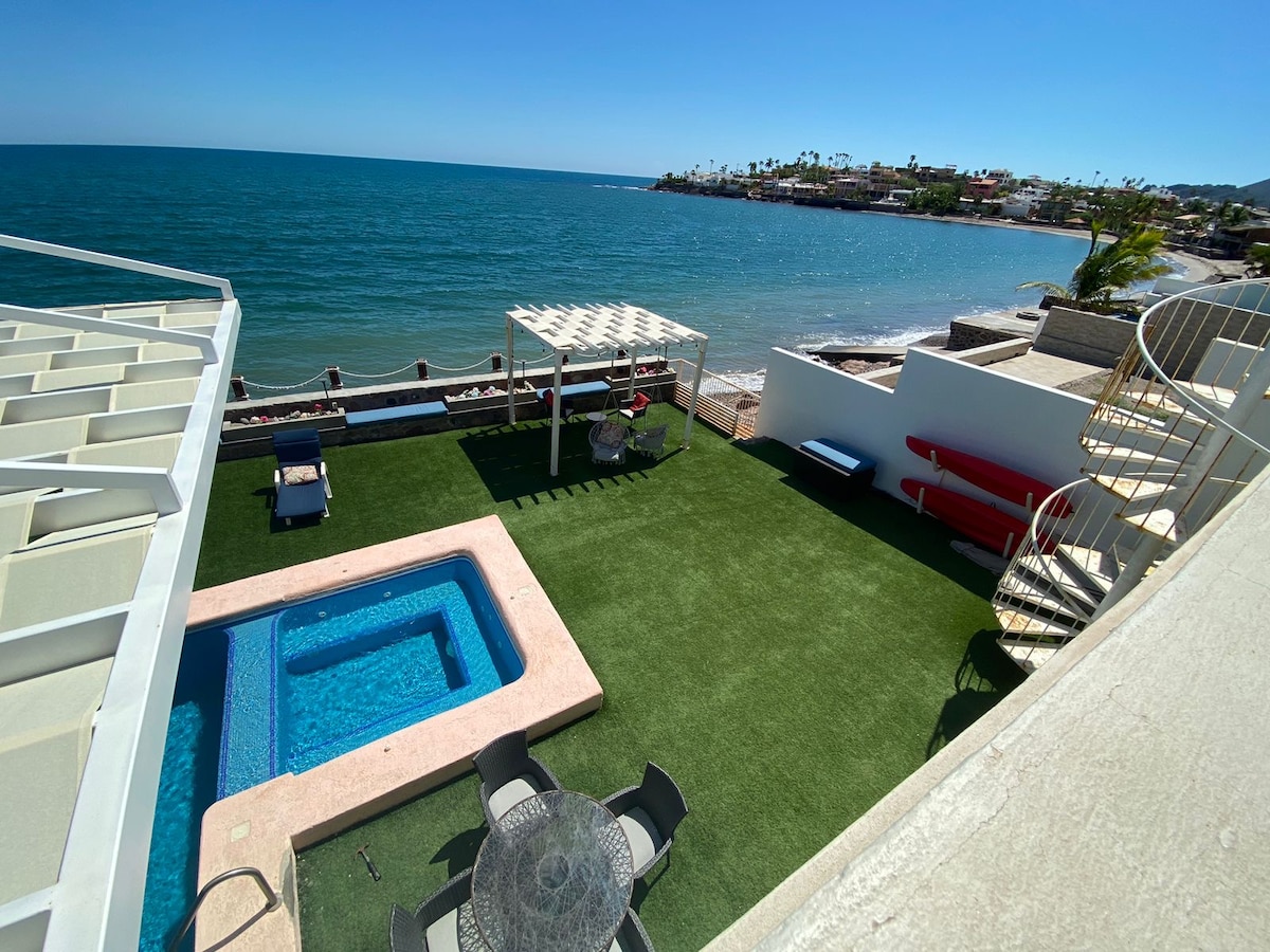 An outdoor space features a private jacuzzi and a pool surrounded by lush green grass. A dining table with chairs and a shaded area are included. The beachfront view showcases calm blue waters and a clear sky, enhancing the serene atmosphere.