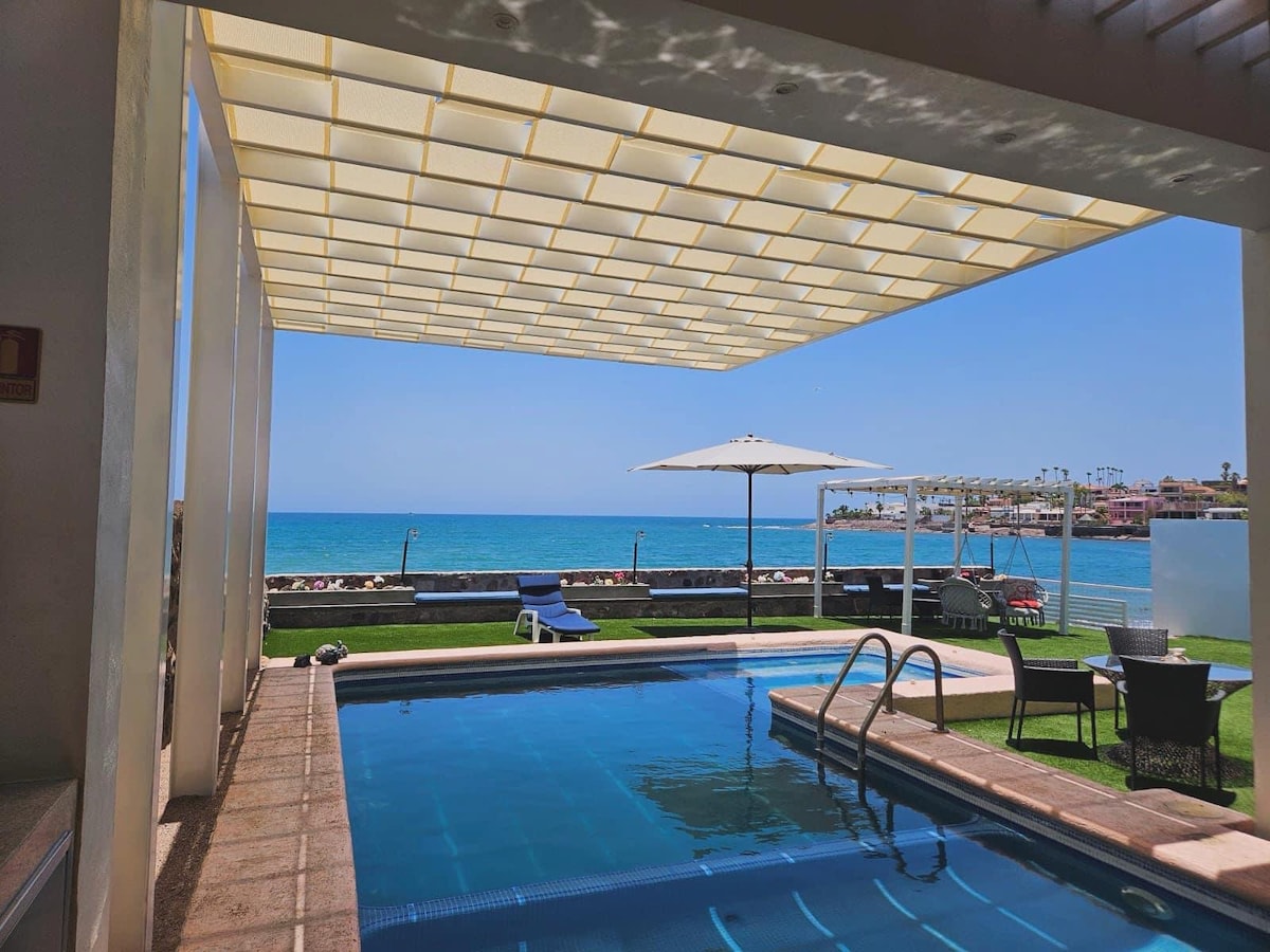An outdoor area features a private pool with a view of the ocean. Lounge chairs are positioned beneath a shaded pergola, and a patio set overlooks the water. Sunlight reflects off the pool, creating a calming atmosphere. The horizon is framed by a clear blue sky.