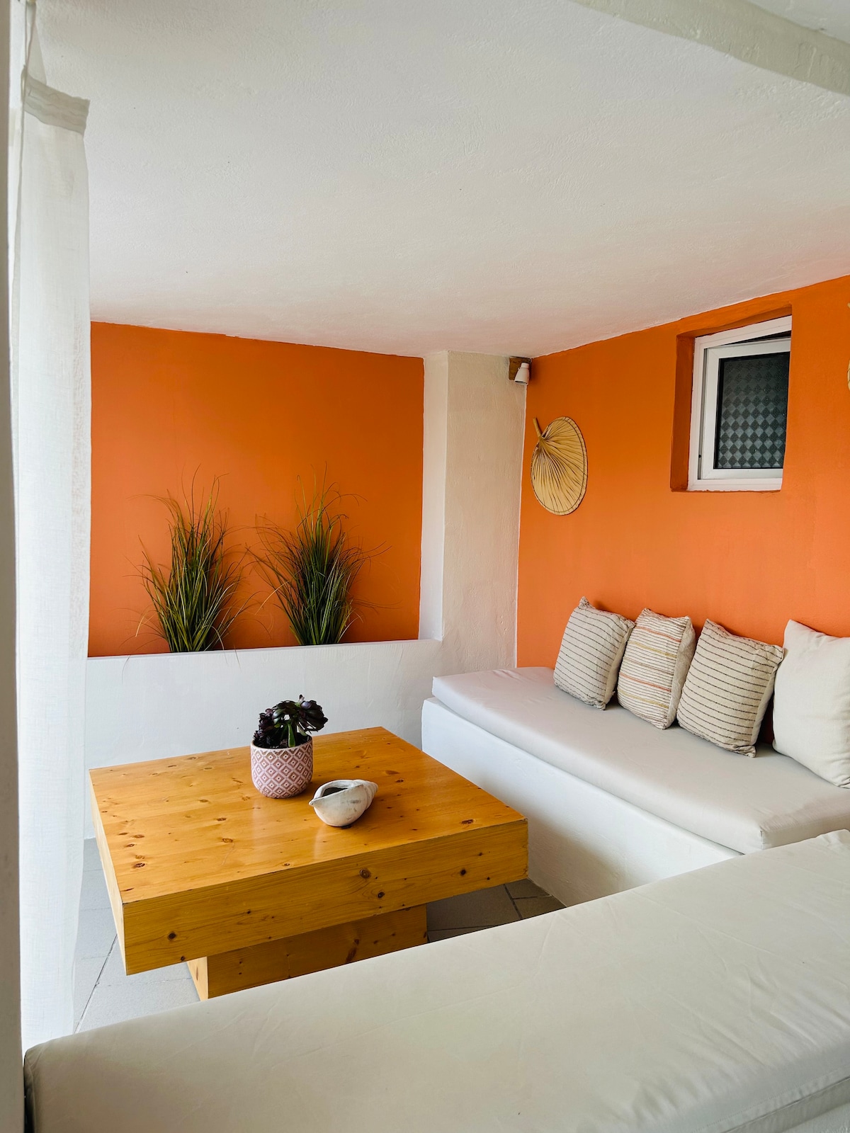 A cozy seating area is nestled against a vibrant orange wall, featuring a wooden table and a collection of neatly arranged cushions. Decorative elements include potted plants and a wall hanging, creating an inviting space for relaxation.