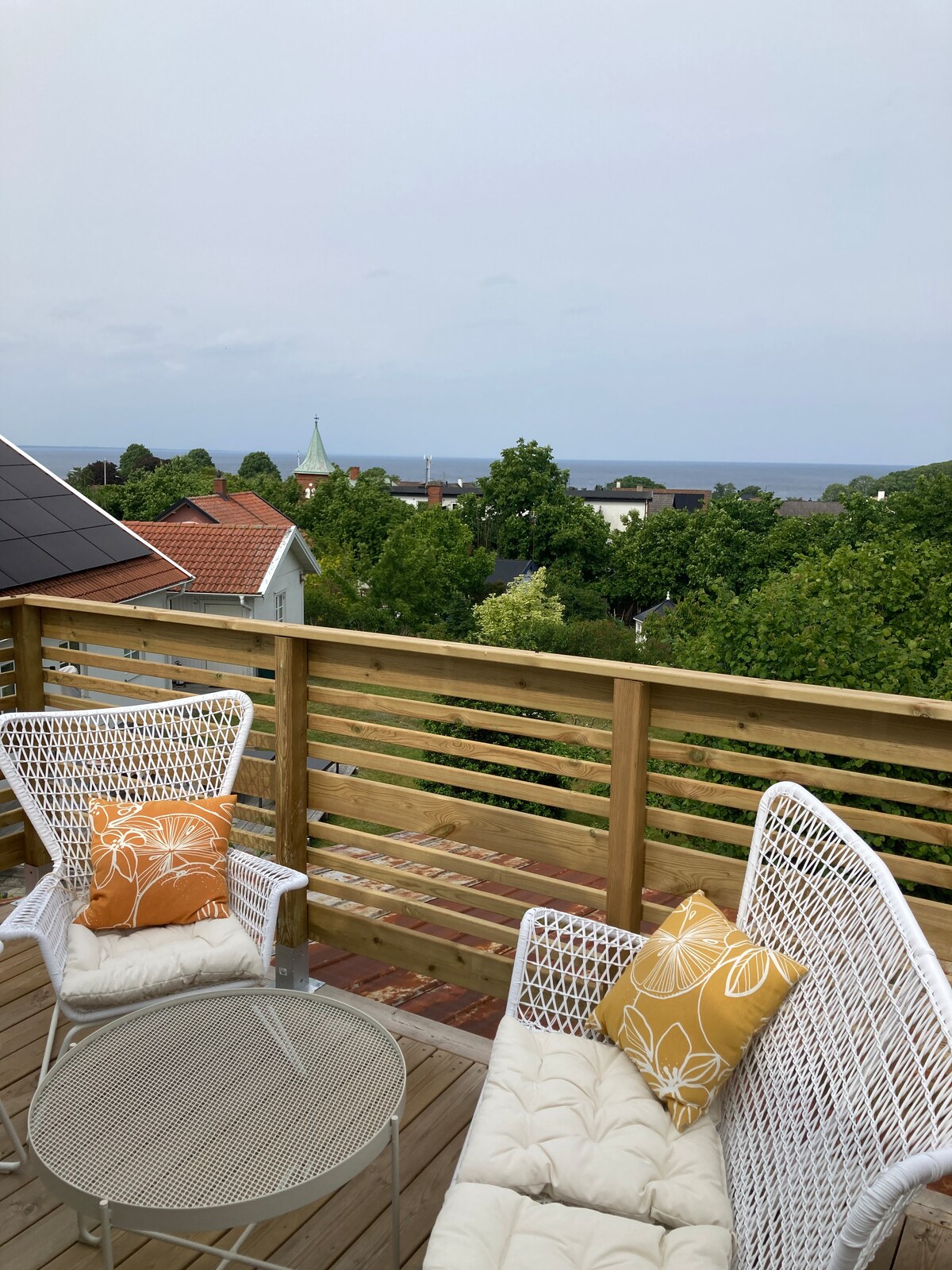 An outdoor balcony furnished with two white wicker chairs and a small glass table. Bright orange pillows add a touch of color. In the background, views of greenery and distant ocean waves can be seen, creating a serene atmosphere.