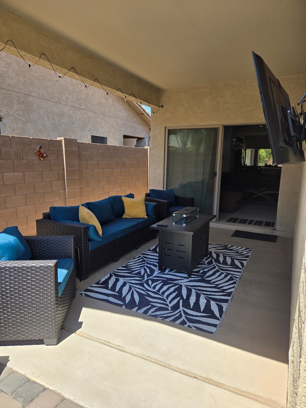 An outdoor living area is showcased with a comfortable seating arrangement featuring dark wicker furniture and bright blue cushions. A coffee table sits on a patterned rug, while a flat-screen TV is mounted on the wall. The space offers a sheltered retreat with a surrounding brick wall.