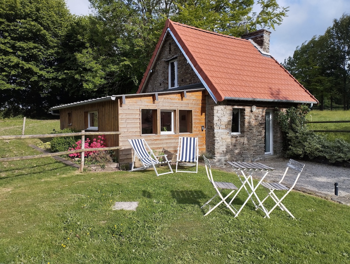 A charming stone cottage is showcased, featuring a red-tiled roof and wooden accents. Two striped lounge chairs and a small white table are situated on the grassy area in front, surrounded by lush greenery and the peaceful countryside.