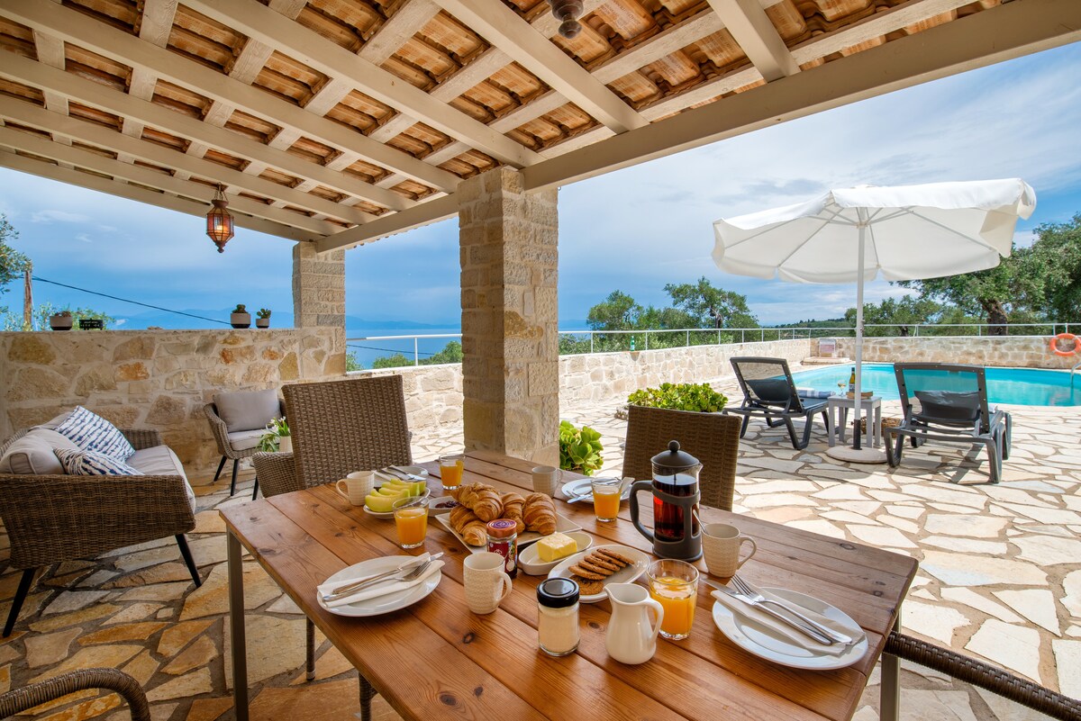 An outdoor dining area is showcased under a covered verandah, featuring a wooden table set with breakfast items including bread, pastries, and beverages. Comfortable seating is arranged nearby, and a swimming pool is visible in the background, framed by lush greenery and a clear sky.