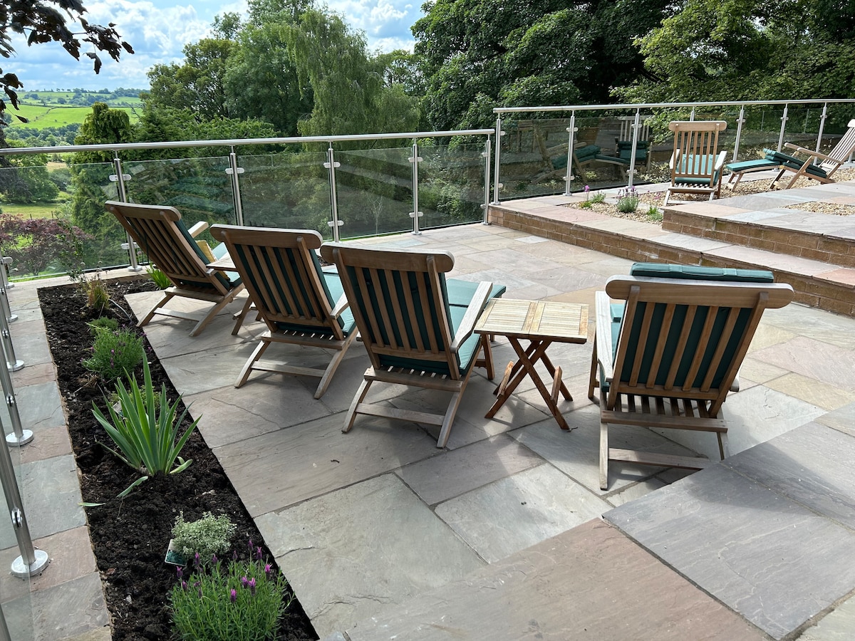 A sun terrace features a set of six wooden steamer chairs with green and white stripes, positioned around a small side table. The area is bordered by a glass balustrade, overlooking the open countryside with greenery in the background.