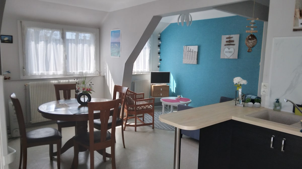 A dining area features a round table surrounded by four wooden chairs. A cozy seating area includes a blue wall, a small television, and a light sofa. Large windows allow natural light to illuminate the space, complemented by decorative items and plants on display.