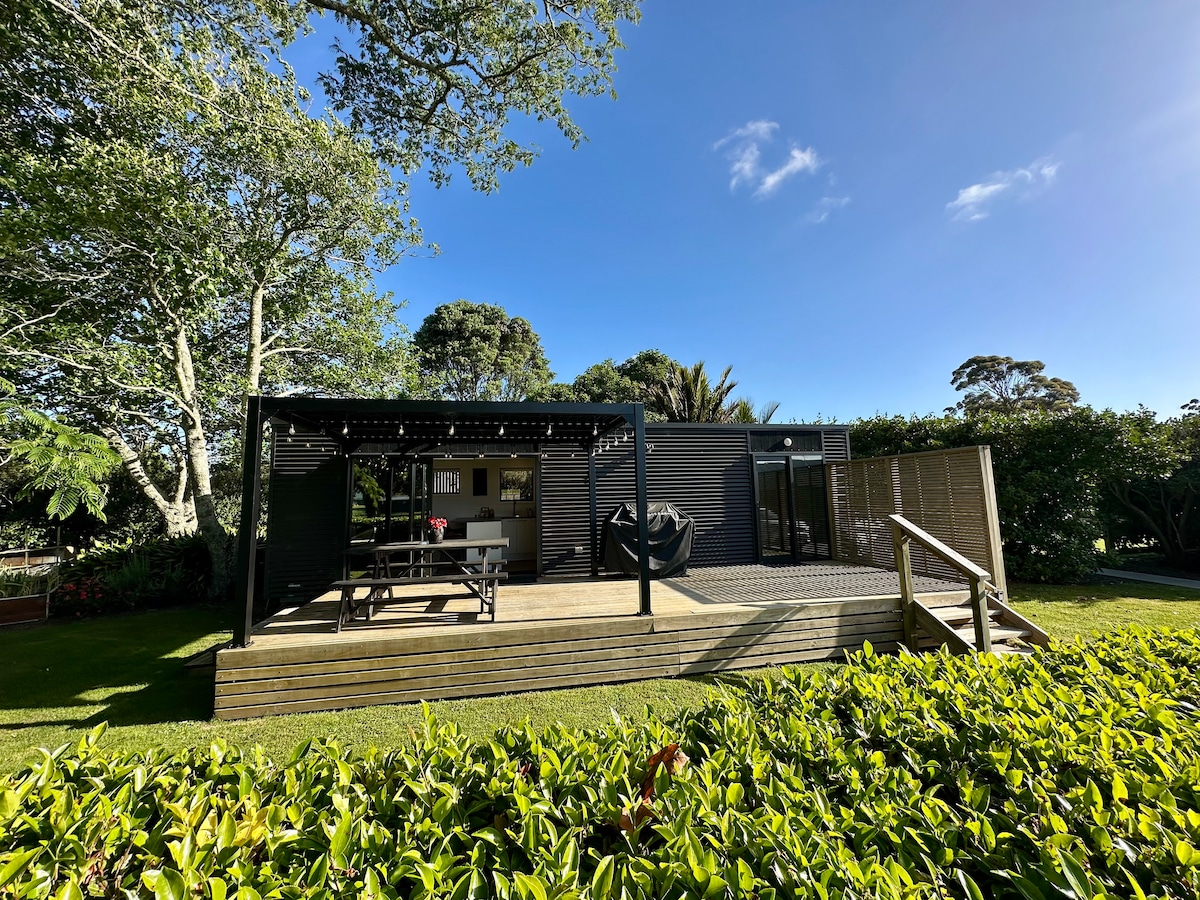 A view of a tiny home surrounded by lush greenery. The deck features a covered dining area with a picnic table, and the entrance showcases large glass doors. Natural sunlight filters through the trees, enhancing the outdoor space's inviting ambiance.