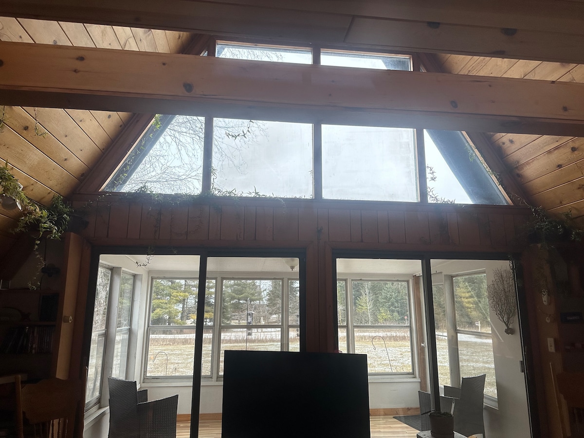 A spacious living area features large triangular windows that allow natural light to fill the room. The wooden beams support the ceiling, while two seating areas with wicker chairs are visible beside expansive glass doors. The serene outdoor view includes trees and an open lawn.