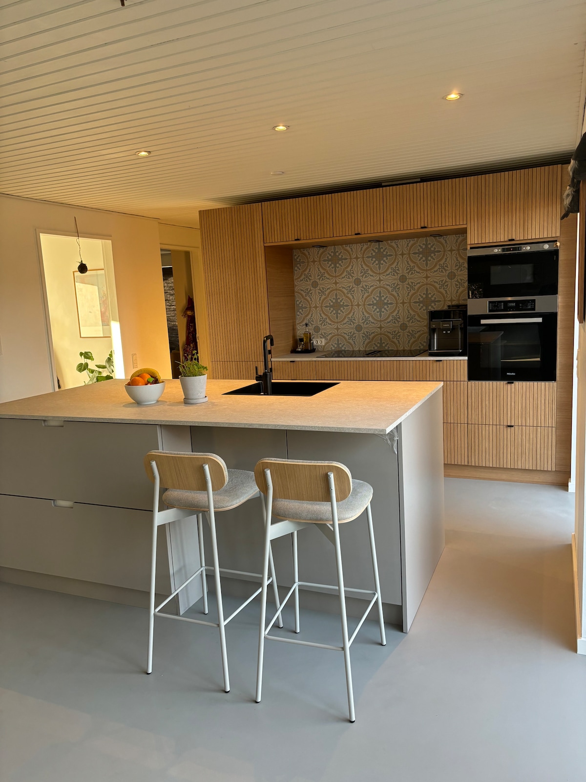 A modern kitchen features a central island with a light grey countertop and two white bar stools. Stylish cabinetry with wood paneling is complemented by a striking backsplash with patterned tiles. Built-in appliances are neatly integrated, and soft lighting enhances the open, airy space.