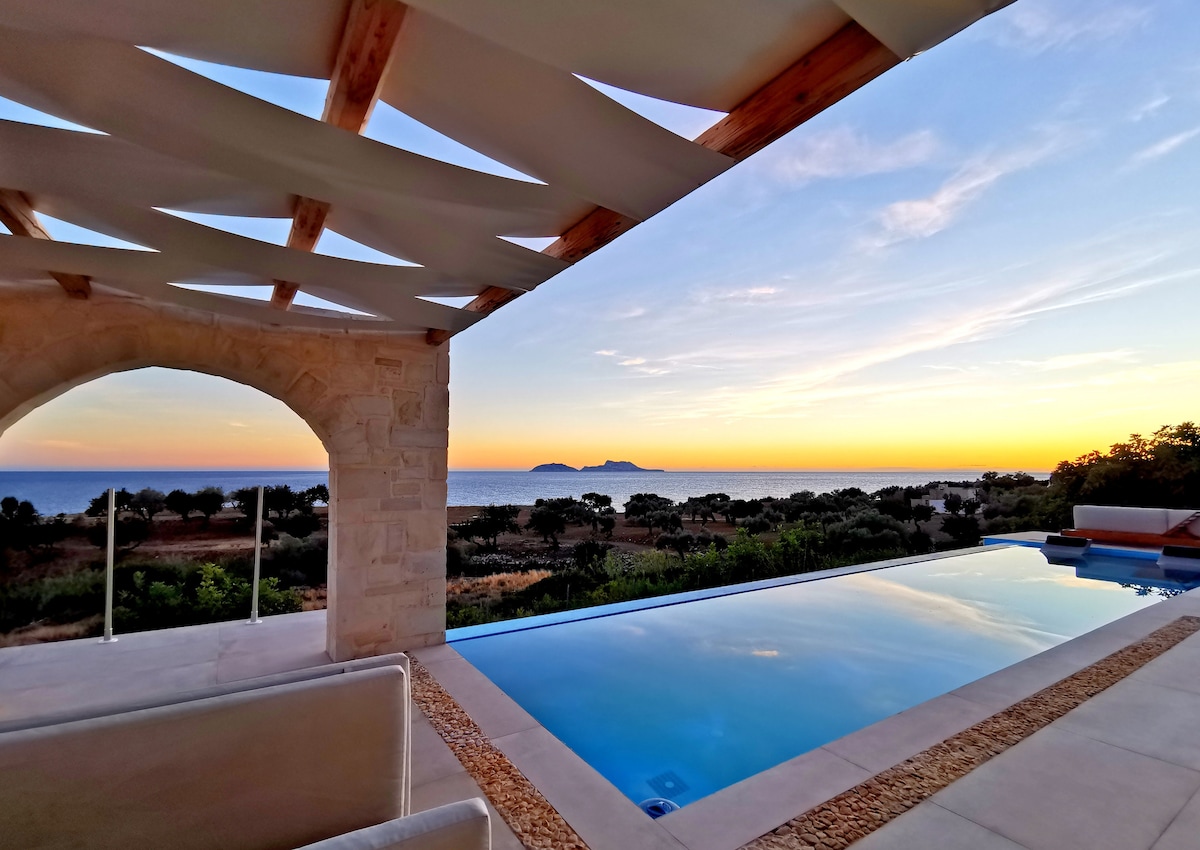A spacious patio is framed by elegant stone architecture, providing a view of the serene sea and islands at sunset. The infinity pool reflects the warm hues of dusk, while olive trees are visible in the foreground, enhancing the tranquil ambiance of the surroundings.