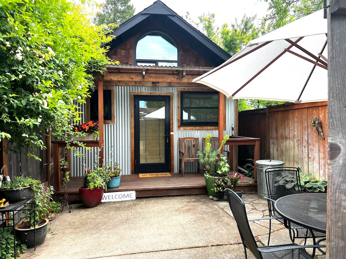 The outdoor porch space is surrounded by greenery, featuring a wooden deck with two chairs and a small table. A large umbrella provides shade, while colorful plants are arranged in pots around the area, creating a welcoming atmosphere.