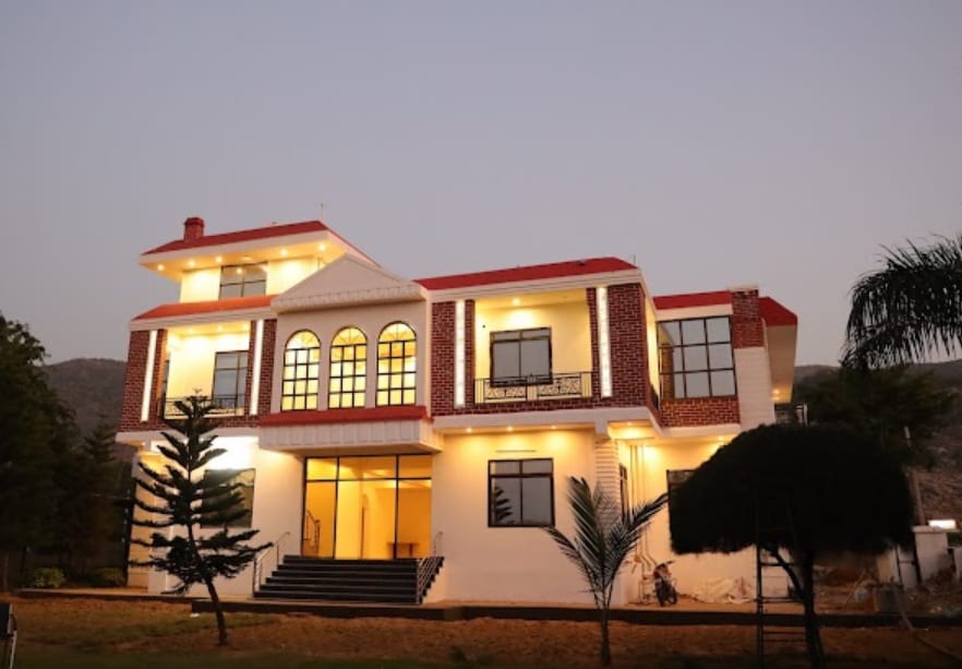 A large, modern house is seen at dusk, featuring a symmetrical facade with a balcony and multiple expansive windows. The building is surrounded by landscaped greenery, while warm outdoor lighting highlights its architectural details.