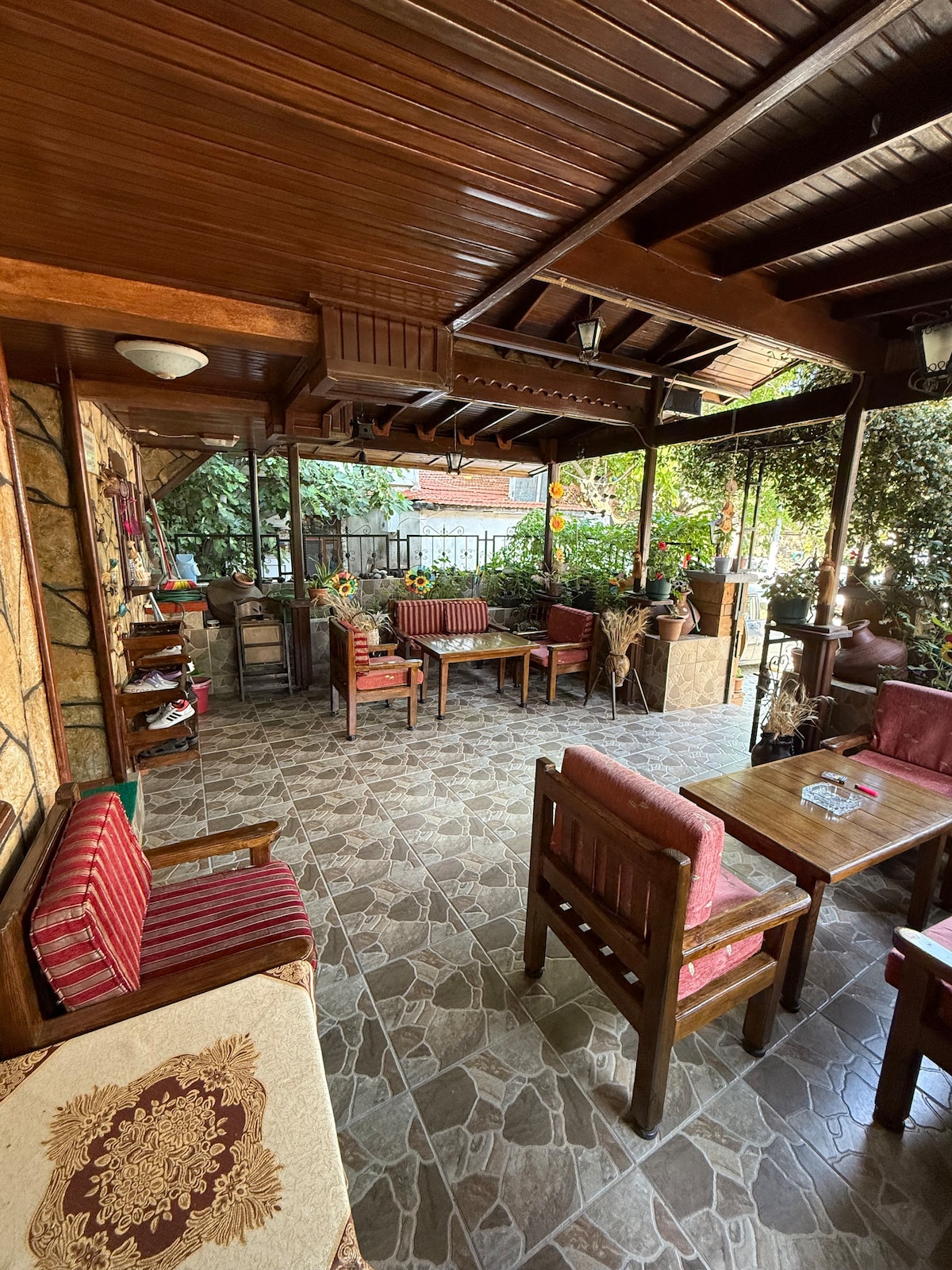 An outdoor seating area features wooden furniture arranged on patterned tile flooring. Plants and foliage provide greenery, while colorful cushions adorn the chairs. Natural light filters through the open space, creating a relaxed atmosphere for gathering.