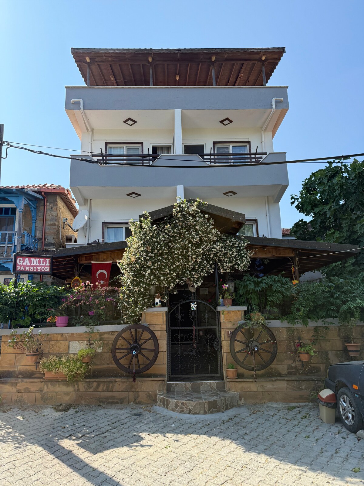 The two-story structure features a modern facade with a spacious balcony on the top floor. A covered entrance area adorned with climbing plants is visible, with decorative elements like wagon wheels enhancing the overall appearance. The surroundings include greenery that adds to the inviting atmosphere.