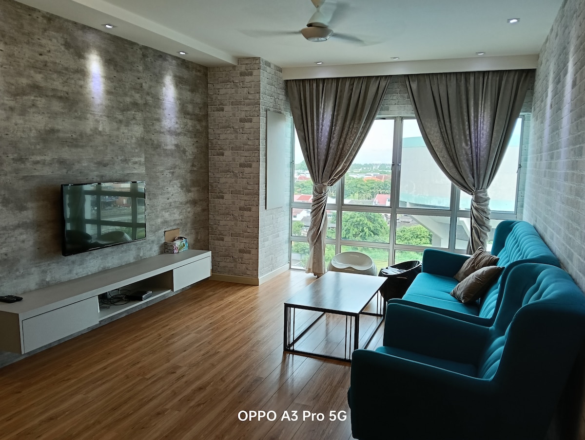 A spacious living area features large windows that allow natural light to fill the room. A comfortable turquoise sofa is positioned next to a wooden coffee table, with a light-toned entertainment unit beneath the wall-mounted television. Stylish curtains frame the windows, adding warmth to the space.