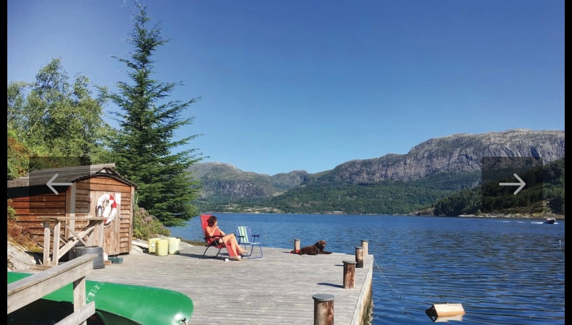 A spacious private dock extends into the calm waters, surrounded by lush greenery and distant mountains. A person relaxes in a chair on the dock, with a dog resting nearby. A canoe is available on the left, offering access to the scenic fjord.