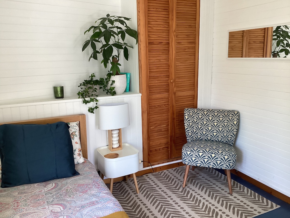A compact sleeping area features a bed with a colorful patterned cover and a dark blue accent pillow. A chair with an intricate design is positioned beside the bed, next to a lamp and decorative plants. A mirrored surface above the chair reflects the warm wooden closet doors.