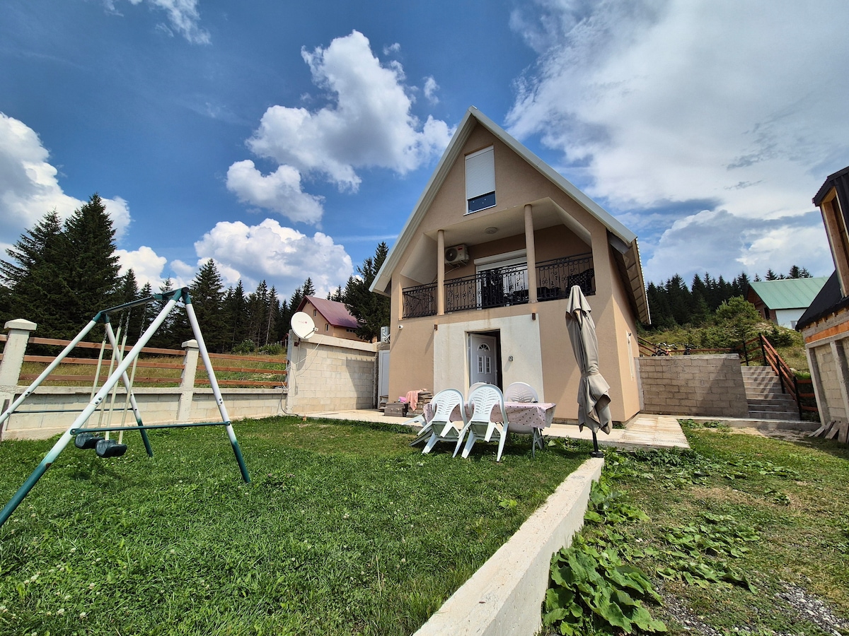 A two-story building features a light-colored exterior and a small balcony. In the foreground, an expansive lawn contains several white chairs and a swing set. The backdrop showcases verdant trees and clouds scattered across a bright sky.