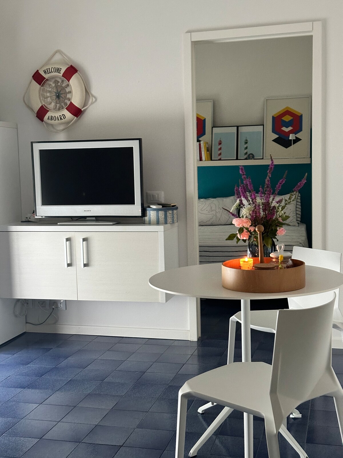 A modern living space is depicted, featuring a white cabinet and a TV on display. A round dining table is set with a decorative centerpiece of flowers and candles. A mirror reflects a cozy sleeping area with colorful artwork visible in the background.