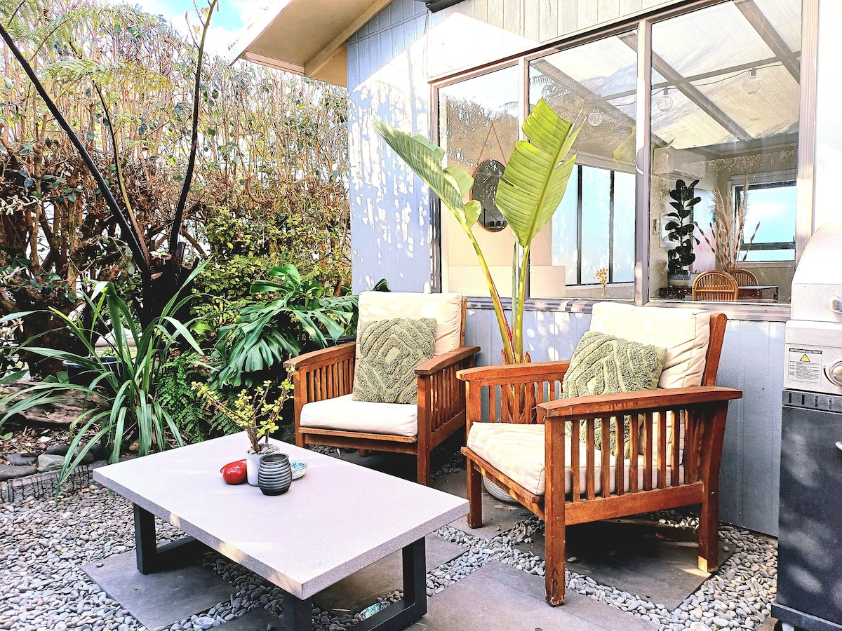 Outdoor seating is provided with two wooden chairs, adorned with decorative cushions. A low table rests between them, featuring a small vase and a few items. Lush greenery surrounds the area, complemented by a large potted plant, creating a serene ambiance for relaxation.