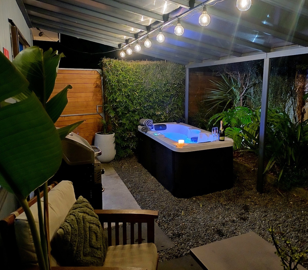 A relaxing outdoor space features a two-person spa pool surrounded by lush greenery and decorative lights overhead. A comfortable seating area is positioned nearby, with a barbecue grill visible on the side.