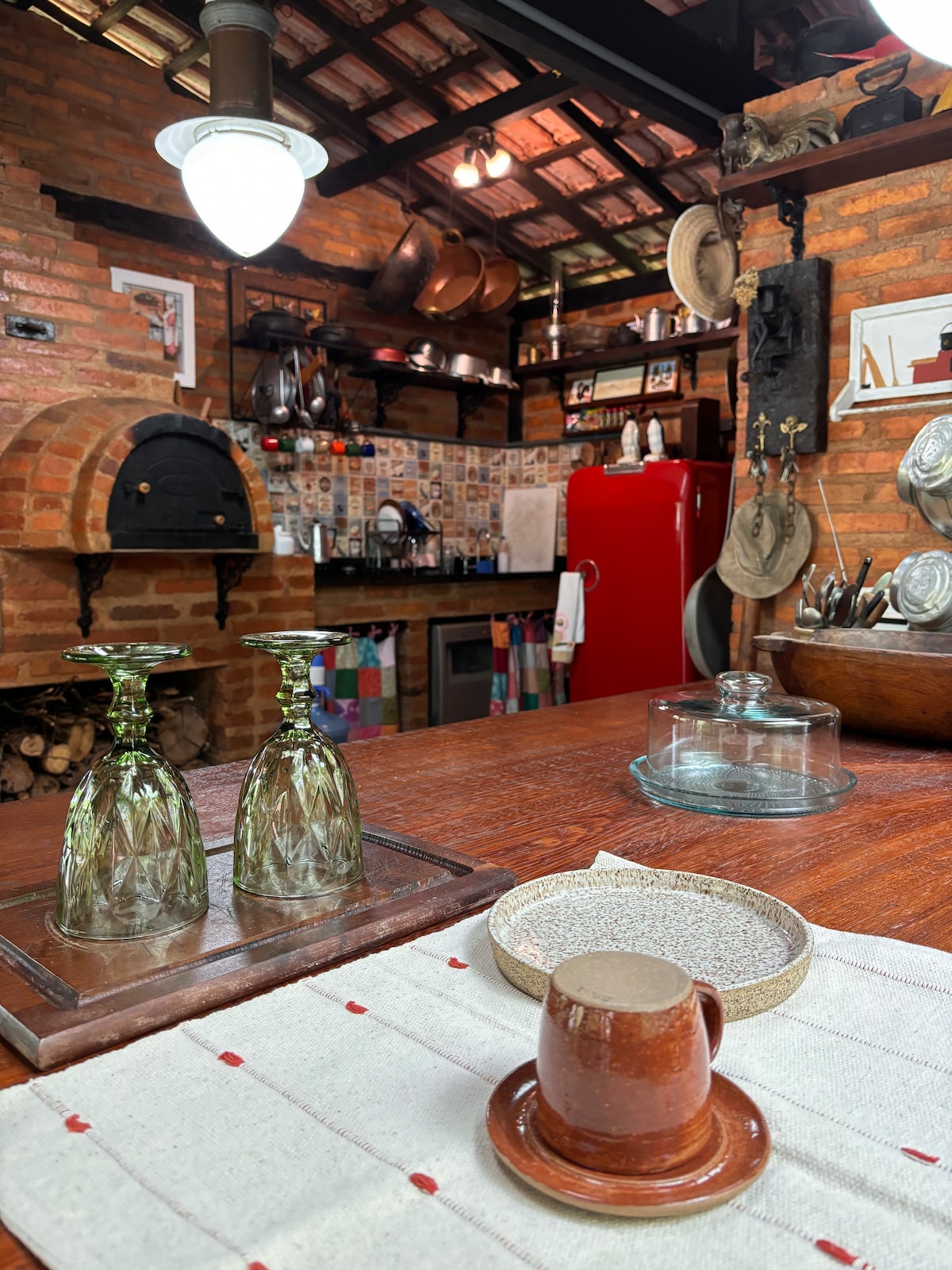 A rustic kitchen is showcased, featuring exposed brick walls and wooden beams. A red refrigerator contrasts with the earthy tones, while glassware and a clay cup are neatly arranged on the wooden table. Various kitchen tools and decorative items add to the charm of the space.