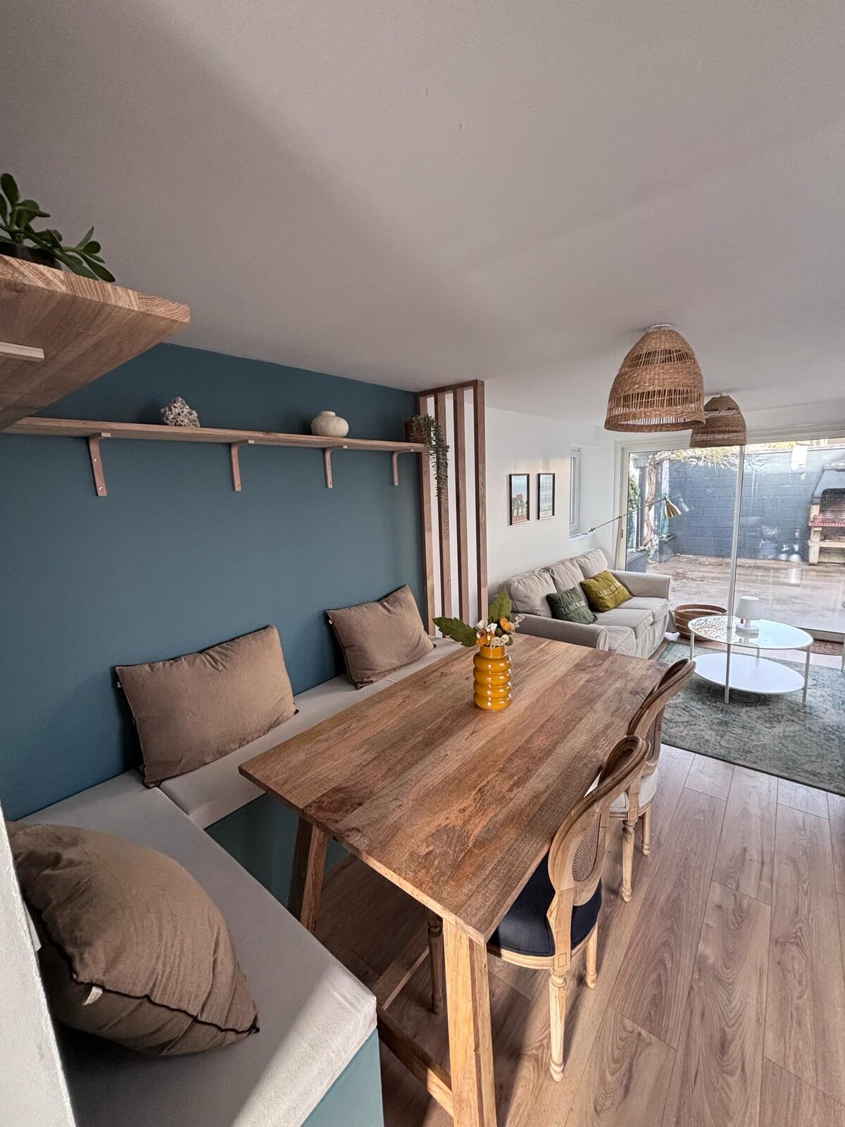 A dining area is highlighted by a wooden table surrounded by chairs. The blue wall serves as a backdrop, featuring decorative shelves with small items. Soft seating areas are present, complemented by natural light flowing from a nearby window, enhancing the inviting space.