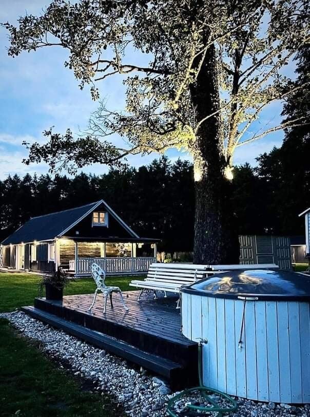 A wooden deck is featured alongside an outdoor hot tub, surrounded by gravel and greenery. A single white chair sits next to a long bench beneath a tall illuminated tree, with a cozy cottage visible in the background, complemented by a starlit sky.