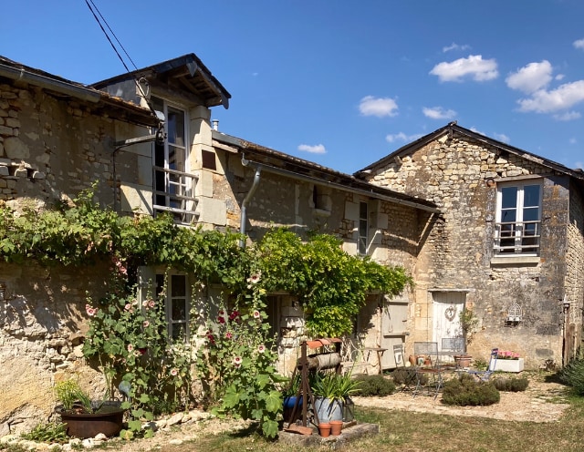 The exterior of the farmhouse showcases rustic stone walls with a vine-covered trellis. A small seating area with metal furniture is featured in the garden, surrounded by greenery. Large windows invite natural light, and the structure is complemented by a clear blue sky.
