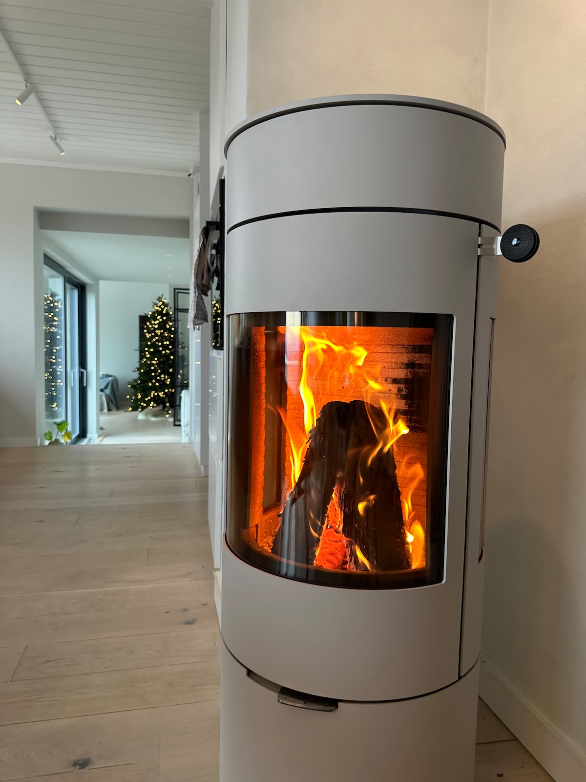 A modern wood-burning stove is positioned in the foreground, showcasing a glowing flame and logs within its clear glass panel. Behind it, an open space leads to a bright area with decorative greenery and a Christmas tree, enhancing the homely atmosphere.