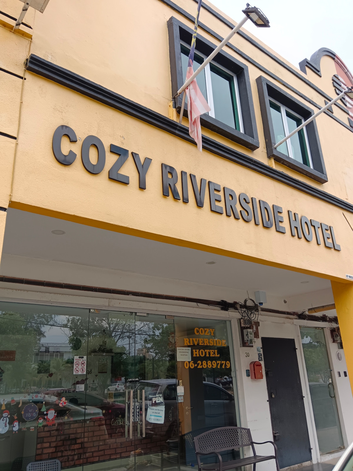 The exterior of a two-story hotel is displayed, featuring a bright yellow facade. The name 'COZY RIVERSIDE HOTEL' is prominently featured in bold letters above the entrance, which includes large windows and a glass door. Two chairs are situated outside, inviting guests to sit.