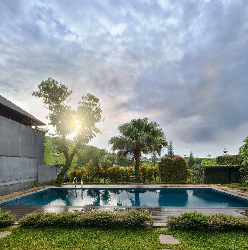 A serene outdoor pool surrounded by lush greenery reflects the sky. Palm trees and well-maintained shrubs frame the space, while a gentle walkway leads to the water's edge. The scene is enhanced by soft afternoon light filtering through the clouds.