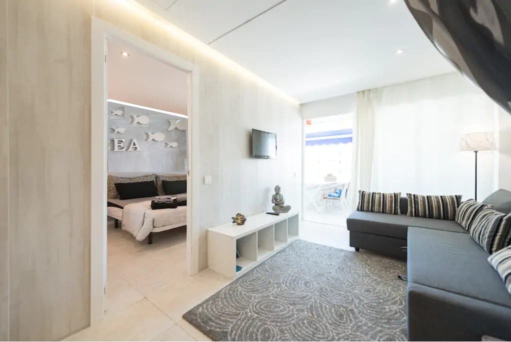 A modern living area features a dark sectional sofa and a decorative rug. A mounted television is positioned on the wall, while natural light enters from large windows. A doorway leads to the bedroom, which includes a soft color palette and nature-inspired decor.