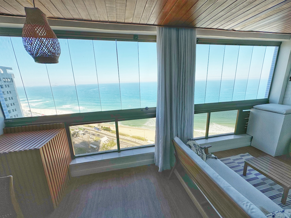 Large windows offer a panoramic view of the ocean, with light filtering through sheer curtains. A stylish seating area is positioned nearby, featuring a wooden cabinet and a cozy sofa, creating a peaceful indoor space that brings the outside in.