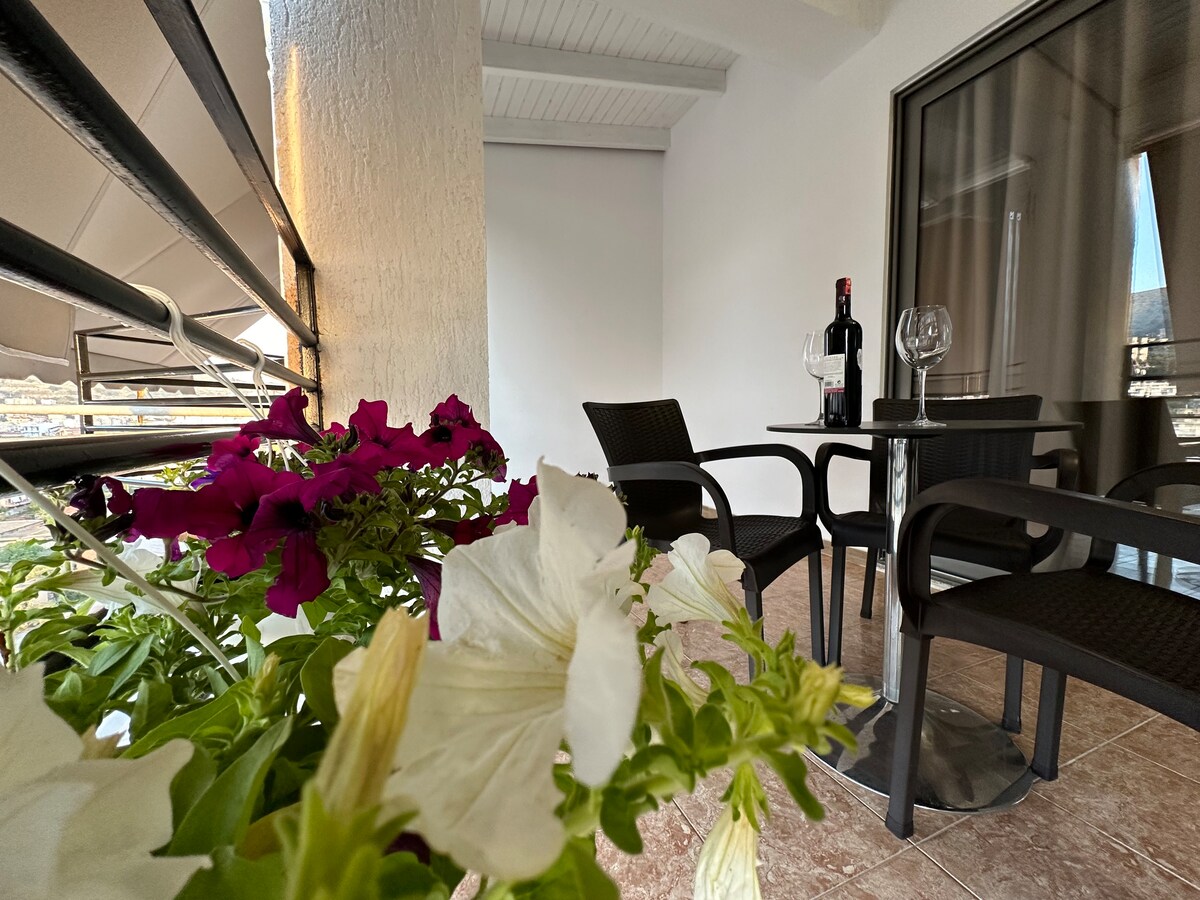 A covered balcony is presented with two dark outdoor chairs and a small table. A bottle of wine and two glasses are arranged on the table. Colorful flowering plants in shades of purple and white add a vibrant touch to the serene outdoor setting.