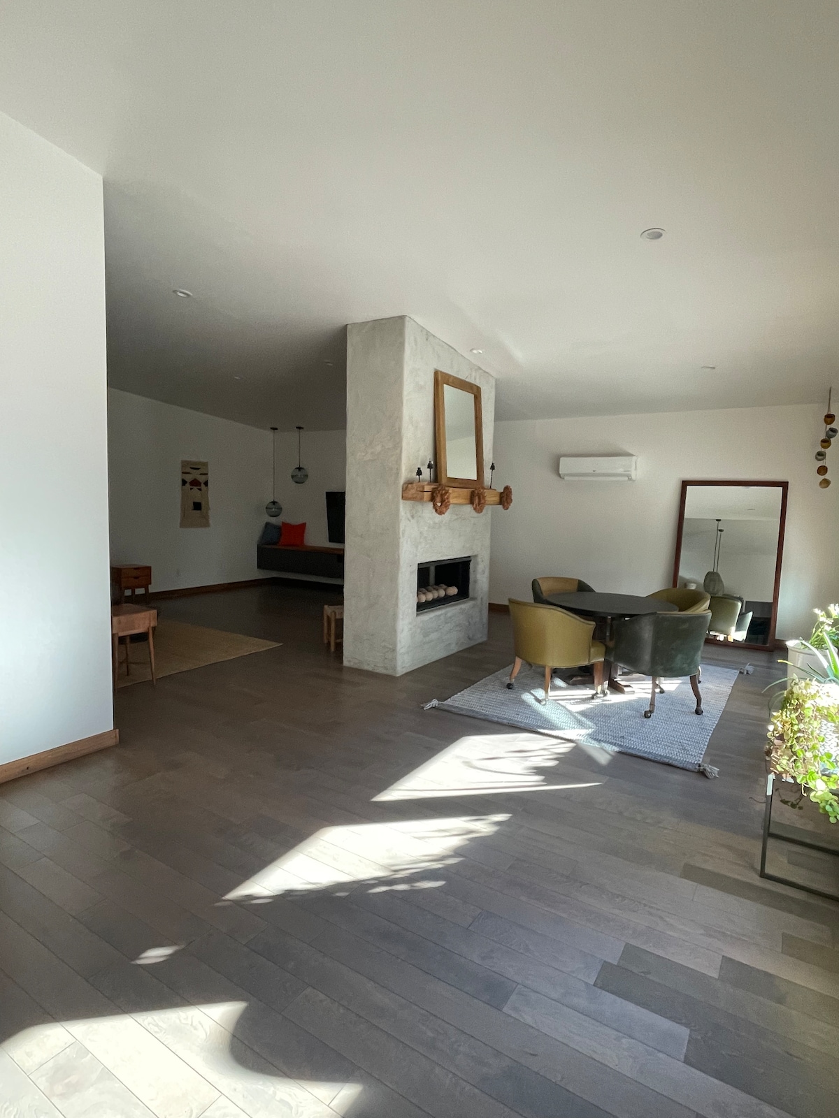 A spacious living area is highlighted by large windows allowing natural light to illuminate the room. A modern fireplace with a decorative mirror above is positioned centrally. The area features a dining table surrounded by chairs and a cozy seating arrangement in the background.